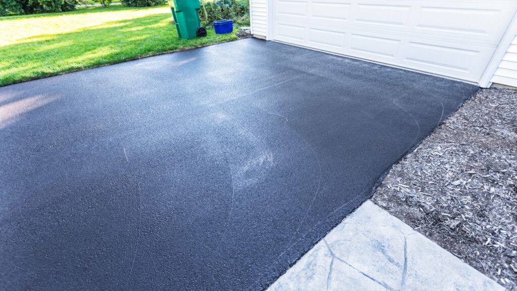 A freshly sealed black asphalt driveway in front of a white garage door, with green grass and a recycling bin visible in the background.