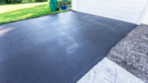 A freshly sealed black asphalt driveway in front of a white garage door, with green grass and a recycling bin visible in the background.