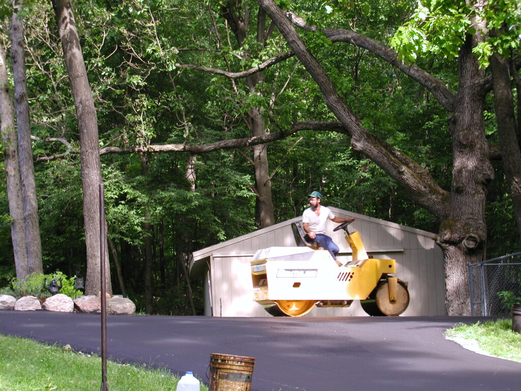 A man operates a yellow road roller to smooth freshly paved asphalt, possibly after foundation repair, in front of a shed surrounded by trees and greenery.