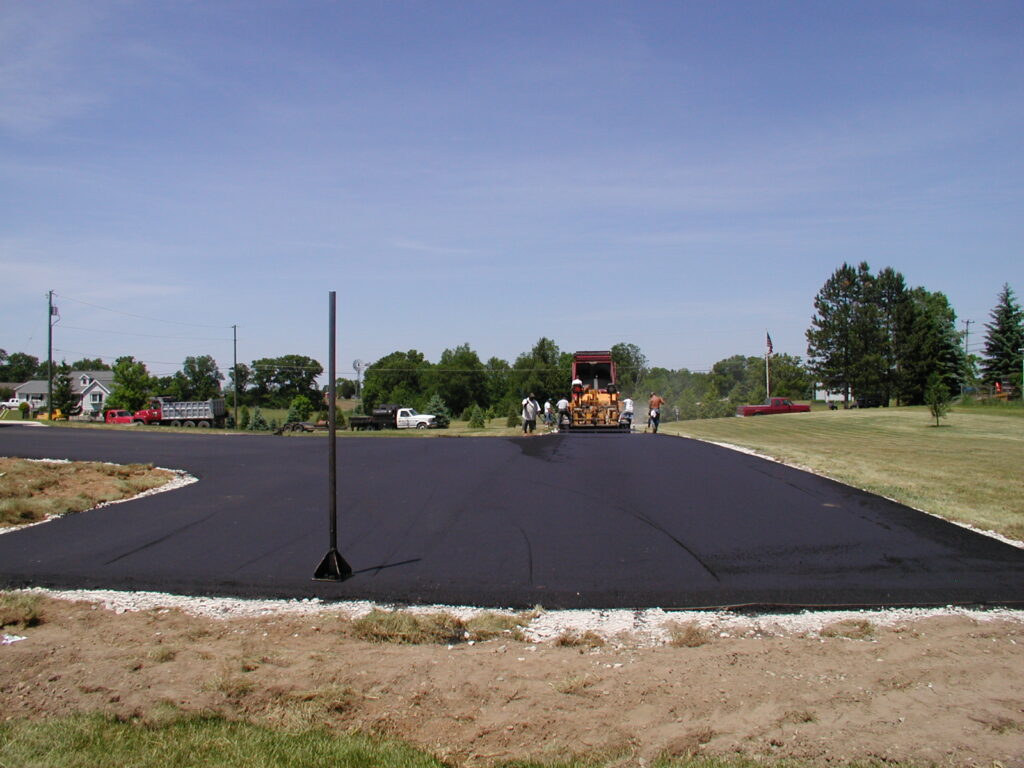 Workers operate paving equipment to lay fresh asphalt on a driveway or parking lot in a suburban area, with trees and a few vehicles in the background.