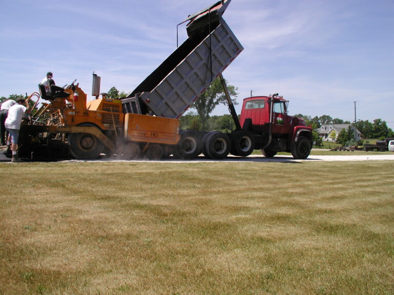 A red dump truck unloads gravel or asphalt into a paving machine while workers are present, preparing a surface near a grassy area on a sunny day.
