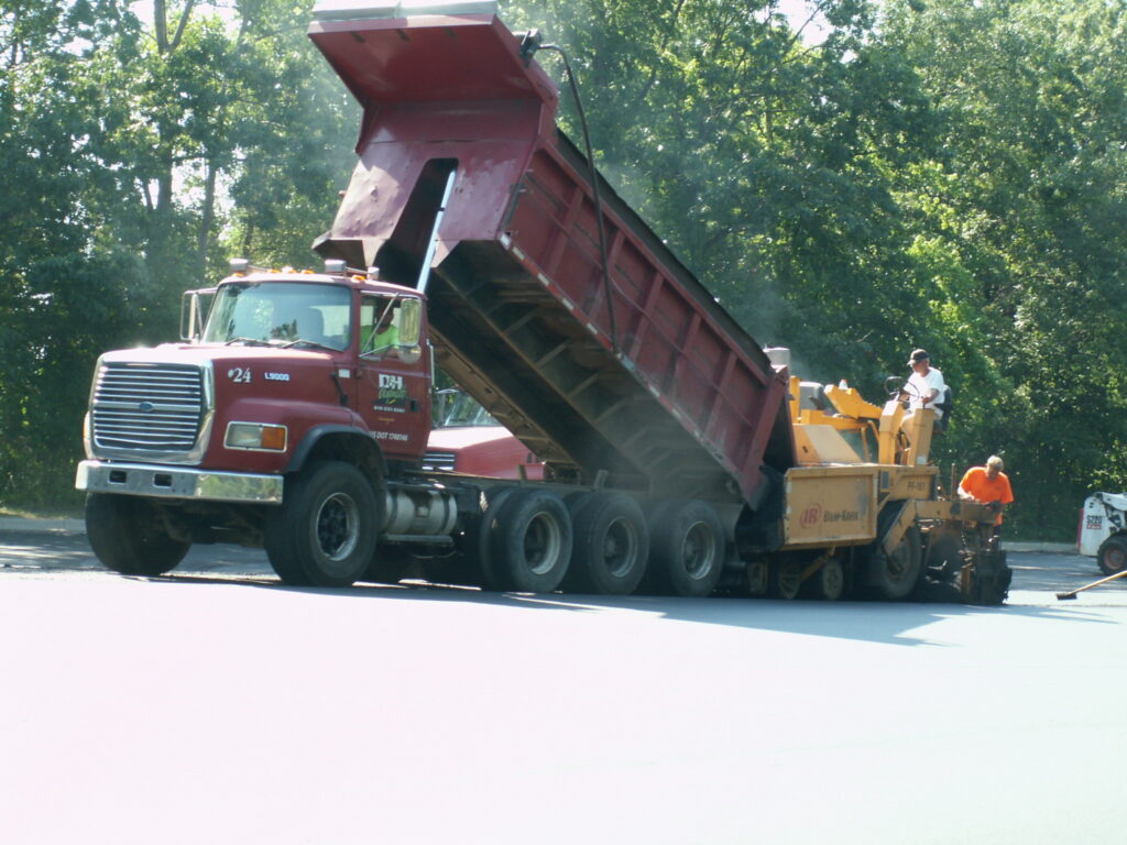 A red dump truck unloads asphalt into a paving machine operated by workers resurfacing a road on a sunny day, ensuring the surface is smooth and strong for future foundation repair needs.