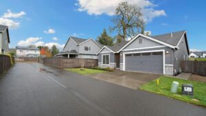 A modern single-story house with gray siding, a double garage, and a fenced backyard, situated along a wet asphalt driveway in a suburban neighborhood.