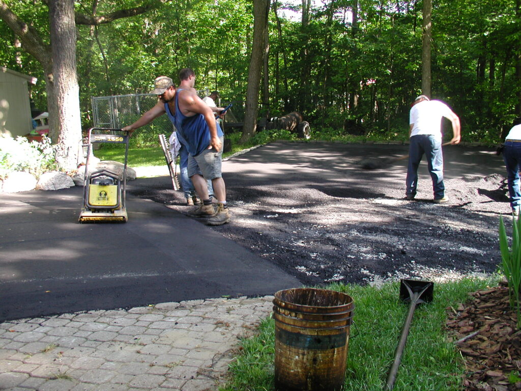 Three men are working outdoors to lay and smooth asphalt on a driveway, following recent foundation repair, using tools and a compactor, with trees and a grassy area in the background.
