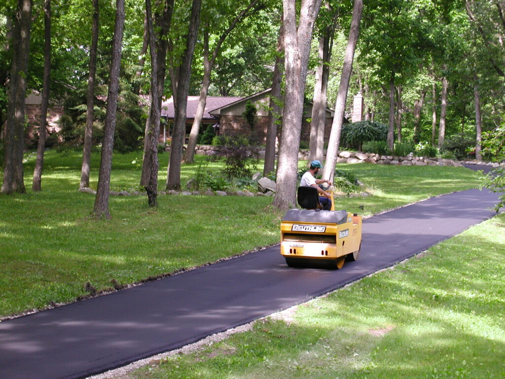 A person operates a yellow road roller on a freshly paved driveway, part of recent foundation repair, surrounded by grass and trees, with a house visible in the background.