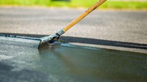 A close-up of a person using a long-handled tool to apply a fresh layer of sealcoating to an asphalt surface.