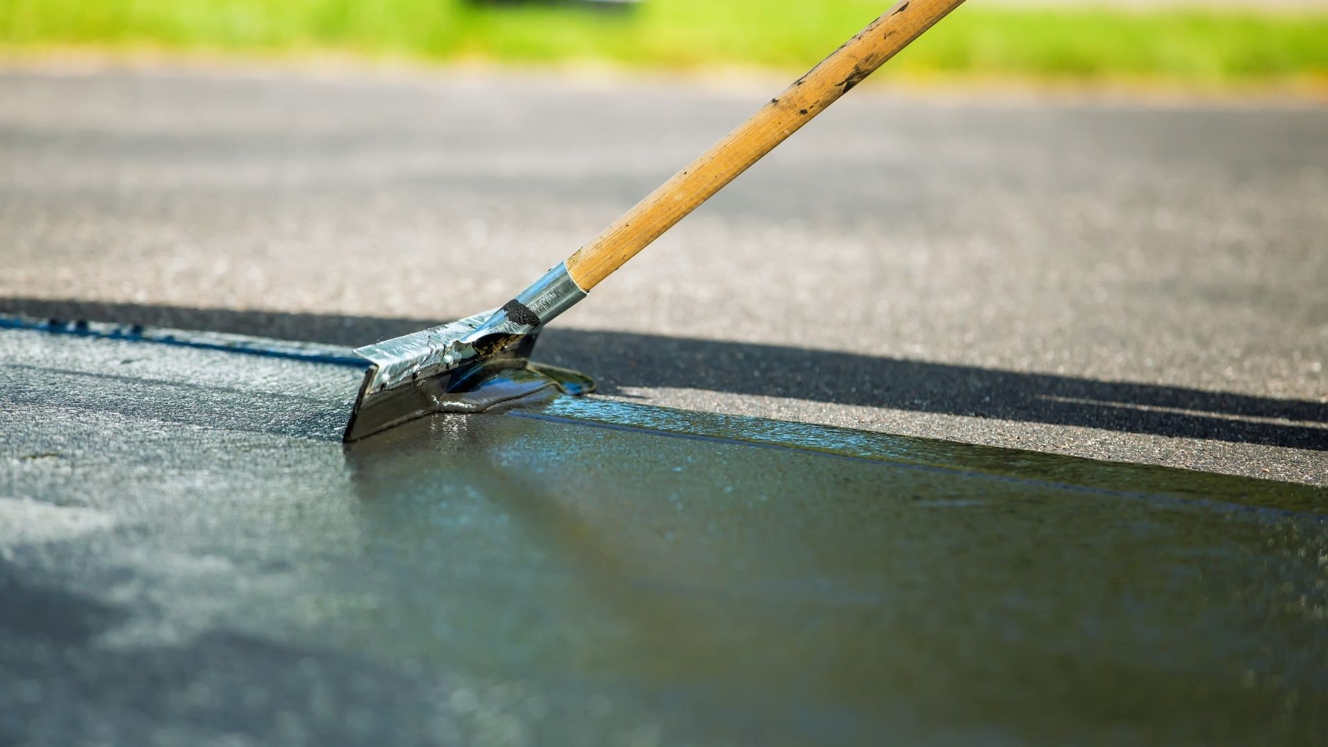 A close-up of a person using a long-handled tool to apply a fresh layer of sealcoating to an asphalt surface.