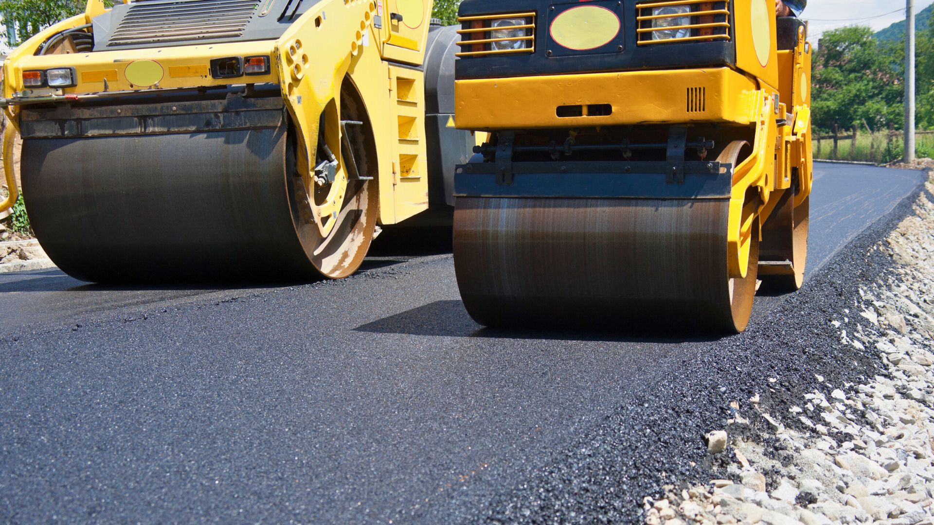 professional asphalt repair in michigan Two yellow steamrollers compact fresh asphalt on a newly paved road under daylight.