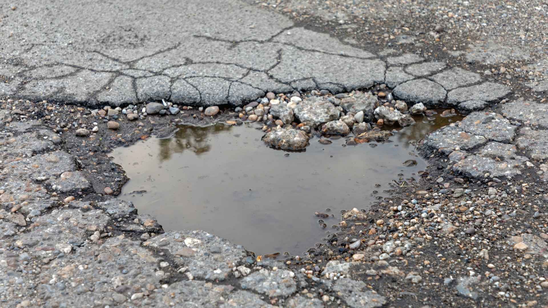 asphalt pothole repair in michigan A pothole filled with water on a cracked, uneven asphalt road.