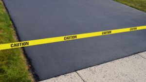 Yellow caution tape blocks access to a newly paved black asphalt driveway bordered by grass and a concrete sidewalk.