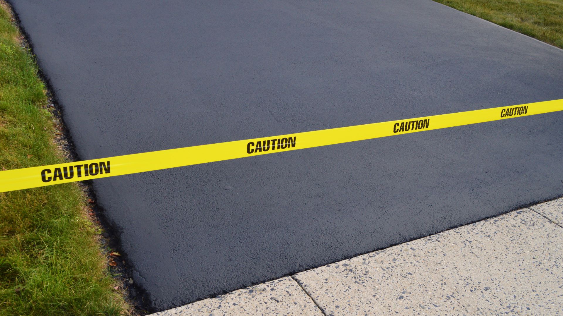 Yellow caution tape blocks access to a newly paved black asphalt driveway bordered by grass and a concrete sidewalk.